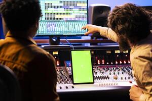 Singer and producer collaborating on new music for an album, looks at greenscreen on tablet next to control panel board in studio. Expert and musician recording new tracks, mixing console. photo