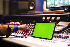 Music producer recording and processing sounds next to greenscreen, using mixing console with knobs and faders. Skilled sound engineer works on technical gear with sliders and colored meters. photo