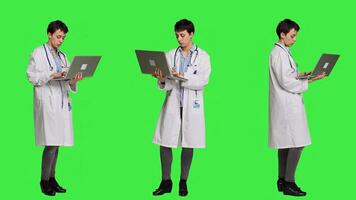 Confident physician searching for new medicaments to treat disease, using laptop to browse on online web pages against greenscreen backdrop. Medic in white coat checks internet sites. Camera A. photo