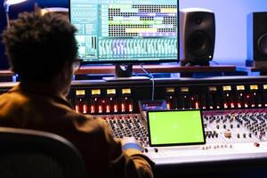African american audio engineer looks at greenscreen on device and digital editing software on computer, processing music in studio. Sound producer creating tracks and adjusting volume levels. photo