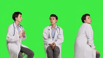 General practitioner acting impatient sitting on a chair against greenscreen backdrop, waiting for patients to attend checkup appointments. Woman specialist in white coat waits for people. Camera B. photo