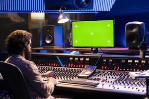 Audio engineer working in studio control room with greenscreen mockup, twisting knobs and switchers to produce music. Producer composer editing tracks and adding sound effects. photo