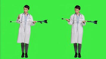Woman physician showing crutches use to help with mobility in case of bone fracture, rehabilitation and recovery after an injury. Medic holds orthopedics tool against greenscreen. Camera A. photo