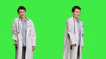 Woman physician showing crutches use to help with mobility in case of bone fracture, rehabilitation and recovery after an injury. Medic holds orthopedics tool against greenscreen. Camera B. photo