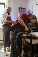 Carpenter and african american coworker using angle grinder on lumber block to smooth surfaces. Man uses orbital sander machinery for furniture assembling, helped by apprentice in background photo
