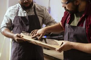 Woodworker holding timber block, showing workshop partner improvements to be made. Manufacturer pointing to apprentice defects in piece of wood to be fixed, close up shot photo