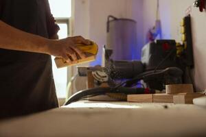 Man using manual sandpaper tool to sander lumber block, assembling furniture in carpentry shop. Carpenter smoothing piece of wood, improving its appearance before applying polish photo