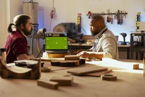 Cabinetmaker looking over blueprints schematics on isolated screen laptop in CAD software with apprentice. Woodworker and colleague designing custom 3D furniture on mockup notebook in assembly shop photo