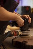 Creative craftsperson using angle grinder on lumber block to smooth surfaces by abrasion with sandpaper. Man using orbital sander gear for furniture assembling work in joinery, close up shot photo