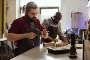 Woodworker next to african american coworker changing power drill head. Carpenter working with BIPOC colleague, preparing to insert screws into wooden surfaces with electric tool photo