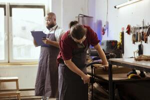Woodworker wearing safety glasses while using bench vise to hold lumber block in workshop next to coworker. Craftsperson using protective equipment and vice tool to clamp piece of wood photo