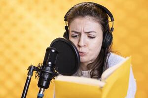 Voice actor with frown on face reading book, recording audiobook using microphone and headset, studio background. Woman using mic to produce digital recording of novel, glowering for dramatic effect photo