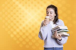 Woman with stack of books in hands enjoying fresh smell of cup of coffee, isolated over studio background. Bookworm holding pile of novels feeling blissful smelling caffeinated drink aroma photo