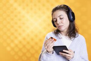 Woman uses ereader to enjoy book, listens music and does purchases online, studio background. Geek reads digital novel on tablet screen, hearing songs in headphones, doing transactions with card photo