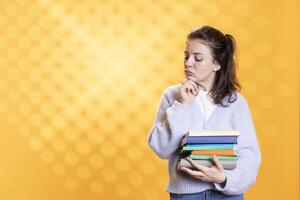 Woman with stack of books in hands deep in thoughts, isolated over studio background. Bookworm holding pile of novels rummaging, thinking about themes and characters after reading photo