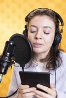 Woman wearing headphones reading aloud from ebook on ereader into mic against yellow background. Voice actor recording audiobook using digital novel on tablet, doing audio story photo