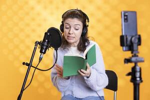 Online star filming vlog at work using smartphone on tripod, reading book to record audiobook. Narrator doing content creation with mobile phone while producing digital recording on novel photo