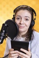 Portrait of radiant woman with headphones reading aloud from ebook on ereader into mic. Voice actor records audiobook using digital novel on tablet, doing audio story, isolated over studio background photo