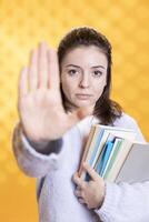 Portrait of annoyed woman doing stop sign while carrying textbooks, tired of preparing for school exam, studio background. Student feeling fed up by university assignments, doing halt hand gesture photo