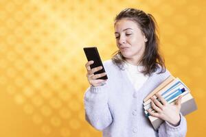 Smiling woman holding pile of books, texting friends on smartphone and enjoying reading hobby. Cheerful lady with stack of novels in arms chatting with mates on phone, studio background photo