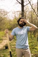 Portrait of overworked exhausted volunteer doing community service, digging holes with a shovel to plant more trees in a forest. Young man activist fighting for reforestation and preservation. photo