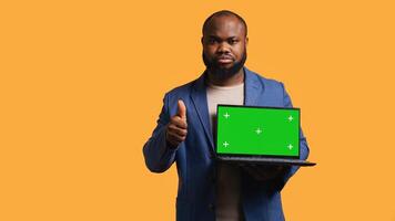 Salesman holding isolated screen laptop, doing recommendation, talking with audience. BIPOC person pointing towards chroma key notebook and showing thumbs up sign, studio background, camera B photo