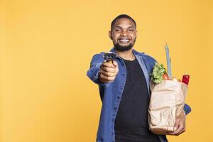 African american young man pointing scanner in front of the camera, scanning various groceries stored in a paper bag against yellow background. Male model with fruits and veggies. photo