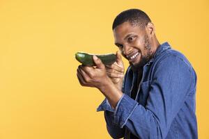 African american funny guy pretending to shoot with a cucumber in studio, playing around and having fun with natural fresh vegetables. Male model acting goofy, posing with raw produce. photo