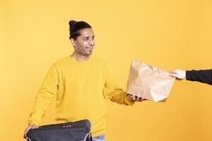 Delivery man arriving to client address with takeaway order in thermal backpack, handing it. Food courier removing packaged meal out of transportation bag, giving it to man, studio background photo