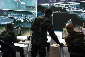 Army recruit conducting an intelligence gathering activity in control room, working on anti terrorism defense plan. Trooper collecting all important data from surveillance footage on big screen. photo