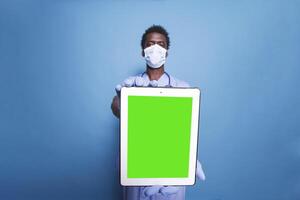 African American healthcare specialist in scrubs and face mask stands with tablet device showing green screen on blue background. Black man holding a tablet with copyspace mockup template. photo