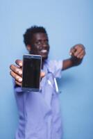 African American physician in scrubs holds mobile device with blank screen. Close-up of male medical nurse with stethoscope grips and gestures to smartphone with empty display. photo