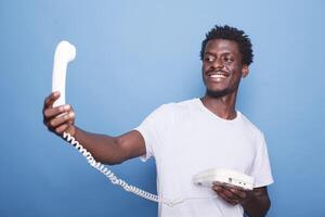 African american gentleman with a smile is holding and looking at the landline phone cord. Black guy with afro standing and grasping a telephone before having a conversation. photo