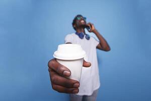 Black man laughing and grasping a coffee cup near the camera. Over an isolated background, a casual African American adult is on the phone, wearing sunglasses, and holding a beverage. photo