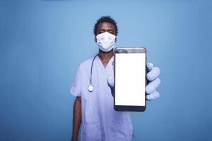 African American man in scrubs grasps a cell phone displaying an isolated white screen. Close-up of male nurse wearing face mask, holding mobile device with blank chromakey mockup template. photo