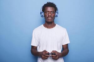 Black man uses headphones and wireless controller while staring at camera. African American guy using joystick to play games, enjoying virtual activities, and listening to music. photo