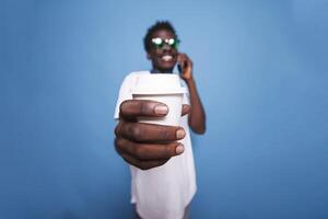 Near the camera, black man is laughing and gripping a coffee cup. Casual African American adult is on the phone over an isolated background, wearing sunglasses and holding a beverage. photo