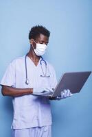 African american man with face mask standing and holding modern laptop. Medical assistant with blue uniform and stethoscope using personal computer in front of isolated background. photo