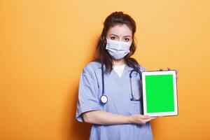 Healthcare specialist vertically holding tablet with green screen display on her left side. Female nurse wearing face mask is gripping a device with blank chromakey copyspace template. photo