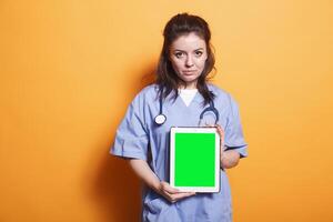 Healthcare professional vertically holding tablet with green screen. Woman wearing nurse uniform and stethoscope is grasping digital device displaying blank chromakey mockup template. photo