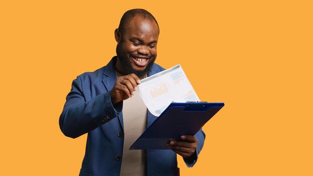 Man browsing though financial audit report on clipboard, pleased by high income, studio background. Happy african american worker reading paperwork on notepad, satisfied by figures, camera A photo