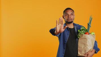Vegan person showing a stop sign with his palm while he carries a bag full of groceries from local farmers market. Young adult restricting the access, expressing rejection in studio. Camera B. photo
