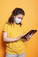 Caucasian lady wearing face mask is holding and using digital tablet for social media. Young woman standing and utilizing modern technology in front of isolated orange background. photo
