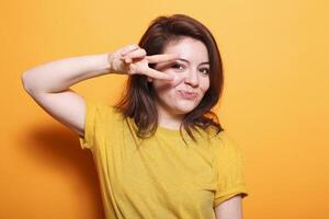 Cheerful brunette with bright smile and casual outfit stands out against isolated background in this waist-up portrait. Carefree lady holding up two fingers to symbolize peace, feeling upbeat. photo