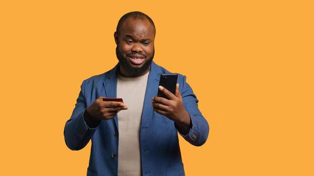 Man upset after lacking necessary money while trying to buy stuff online on smartphone. Sad person looking at debit card after getting transaction rejected on phone, studio background, camera A photo