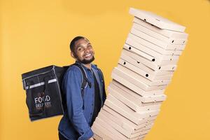 Smiling deliveryman struggles to hold a huge stack of pizza boxes in studio, working on food delivery service to bring orders to clients. Express courier with a backpack carries stack of takeout. photo