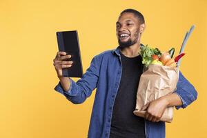 Smiling young man checks something on his tablet while he is holding a bag full of fresh vegetables and groceries. Confident guy uses his mobile device for social media against yellow backdrop. photo