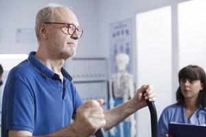 Side-view portrait of caucasian elderly man using stationary bicycle for physiotherapy with assistance from female nurse in blue scrubs. Senior patient in hospital for rehabilitation recovery. photo