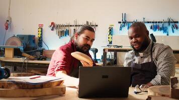 Carpenter and colleague verifying finished wood art product, comparing with blueprint. Artisan and apprentice inspecting wooden object for damages based on schematics on laptop screen, camera B photo