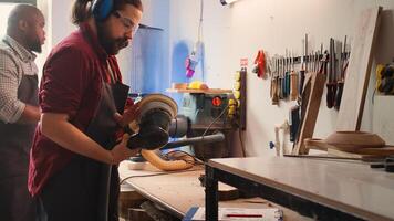 Carpenter at work consulting blueprint, using orbital sander on lumber, wearing safety glasses. Artisan with protecting equipment uses angle grinder on wood after checking schematics, camera B photo